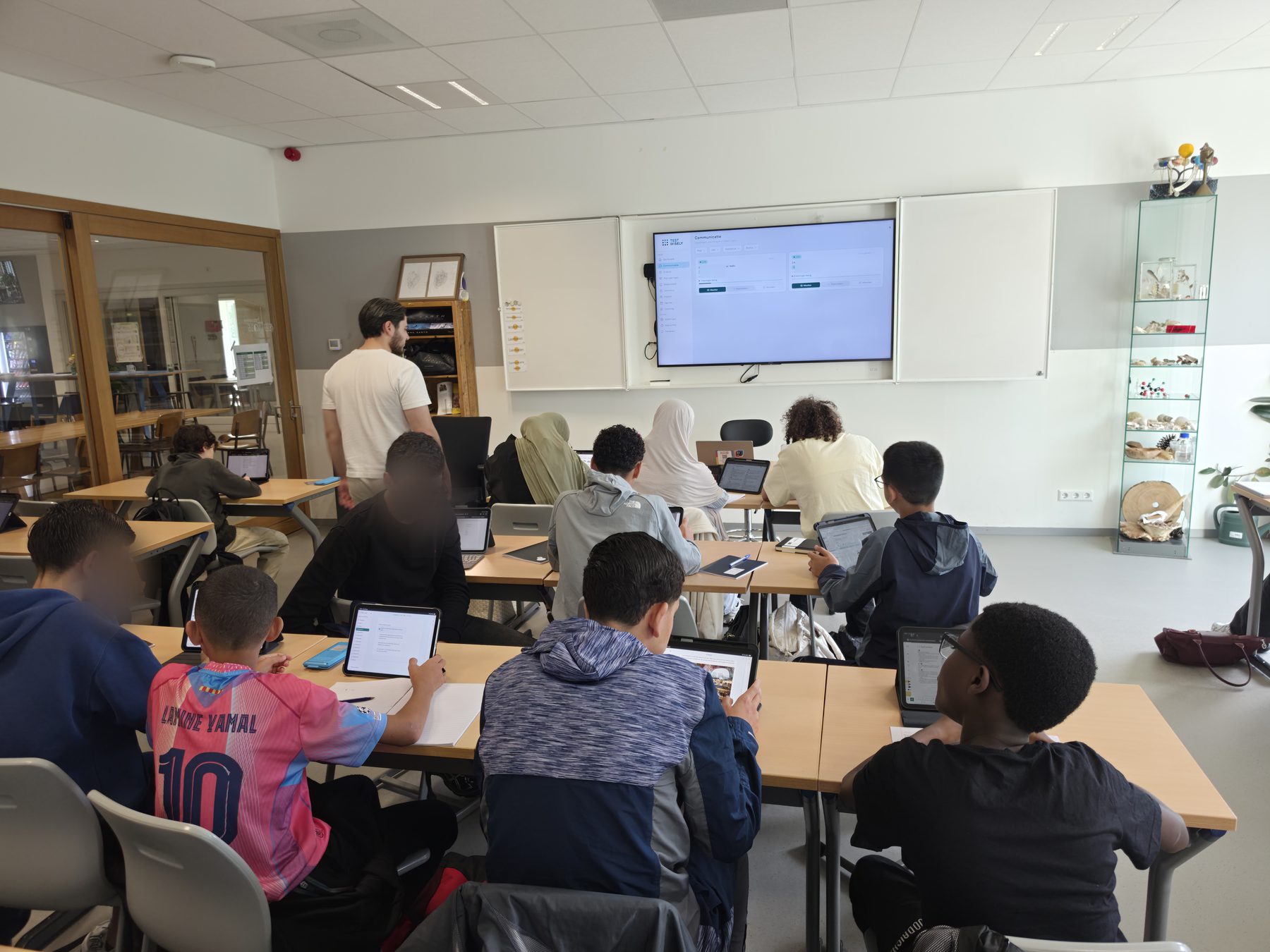 Leerlingen werken op iPads in TestWisely tijdens een les; op het scherm vooraan staat het communicatie-overzicht.